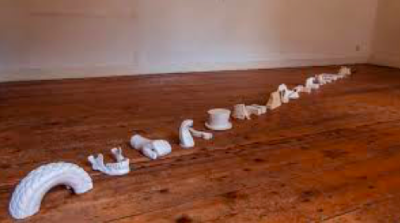 De witte sculpturen gebaseerd op fotoherkenning liggen diagonaal op een houten vloer van links naar rechts. Eerst zie je een halve autobandm dan een onderkaak en andere willekeurige voorwerpen.