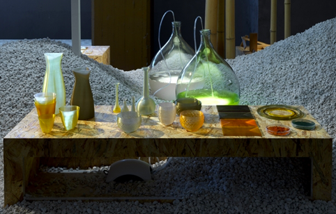 Een installatie van Atelier Luma met glazen kolven gevuld met algen, omringd door proefbuisjes, potten en designobjecten op een tafel van gerecycled materiaal. Illustratie van kunst en duurzaamheid met algen.