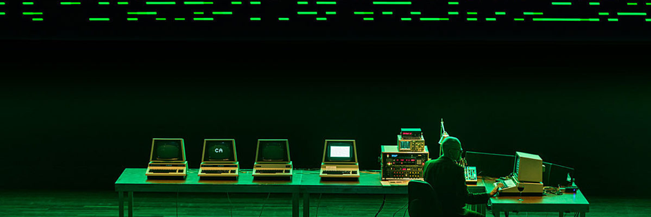 De verschillende computers op een groene tafel, waar een man aan zit te werken. De computers werken samen om een digitaal kunstwerk te projecteren in zwart en groene pixels.