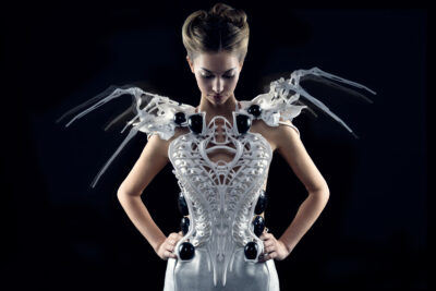 Een vrouw draagt de futuristische "Spider Dress" ontworpen door Anouk Wipprecht. De jurk is een wit, 3D-geprint exoskelet met ingewikkelde patronen en zwarte accenten. Mechanische, spinachtige structuren strekken zich uit vanaf de schouders. De achtergrond is donker, wat de high-tech en avant-gardistische uitstraling van de jurk benadrukt.