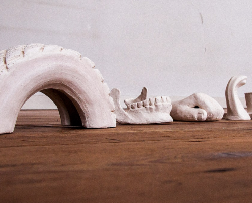 Witte sculpturen gebaseerd op fotoherkenning liggen in een rijtje op een houten vloer. V.l.n.r. halve autoband, onderkaak, hand.