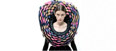 Model draagt Behnaz Farahi’s Iridescence, een opvallende ketting met kleurrijke, visachtige schubben die reageren op beweging. Deze interactieve mode benadrukt technologie en innovatie.
